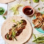 Hoisin chicken wraps laid out on a wooden board with cucumber matchsticks, shredded lettuce, spring onions and a bowl of hoisin sauce